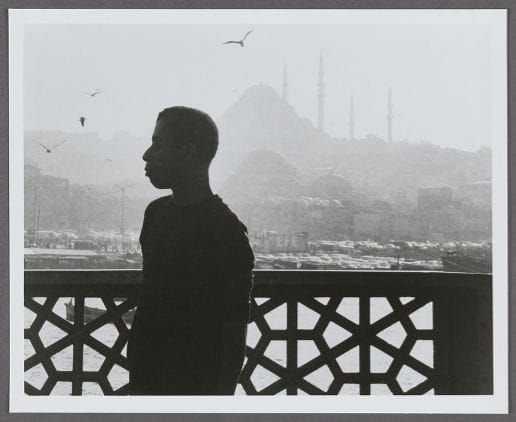 Black-and-white photograph of writer James Baldwin in dark profile silhouette, gazing left toward a hazy skyline dotted with minarets and ornate geometric railing in view.