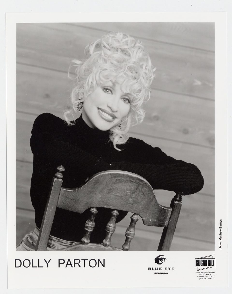 Black-and-white portrait of Dolly Parton smiling, with voluminous blonde curls, leaning on a wooden chair.
