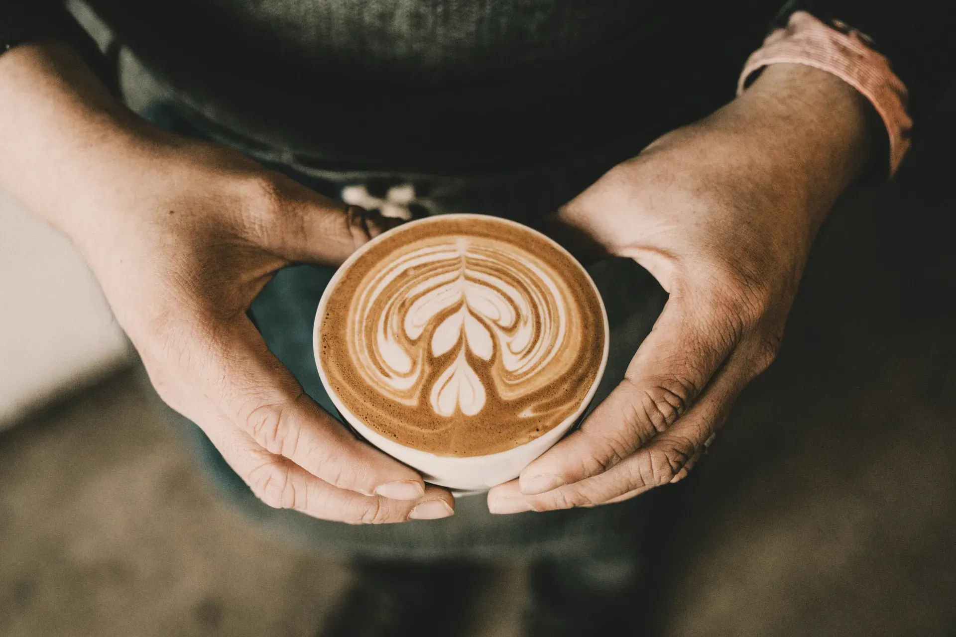 Two weathered hands cupping a latte with latte art. The hands show signs of work and wear. The coffee looks perfect. The person behind it remains unseen.