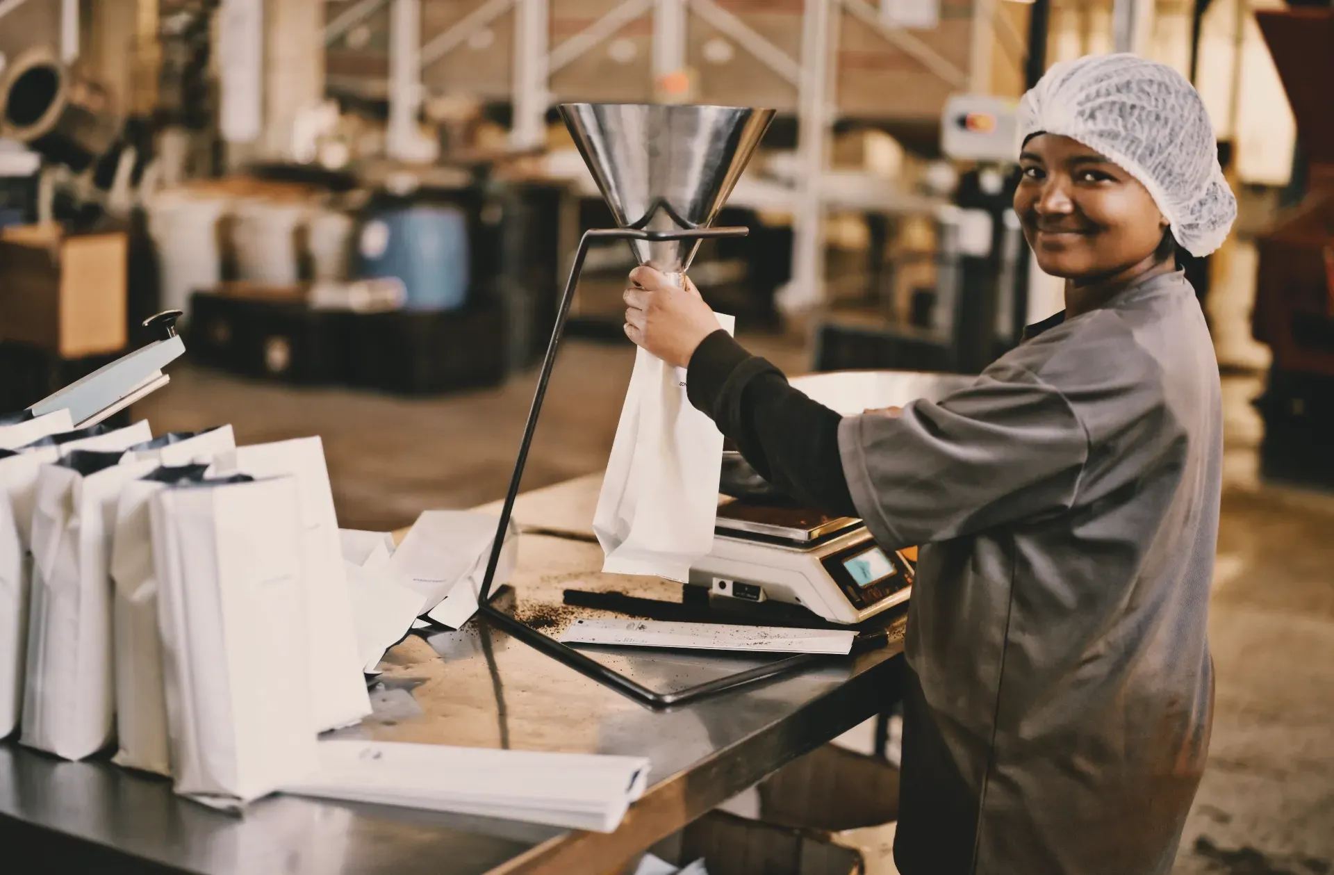 A woman in a hairnet and work apron fills coffee bags at a packaging station in a roastery. She is smiling and looking toward the camera 