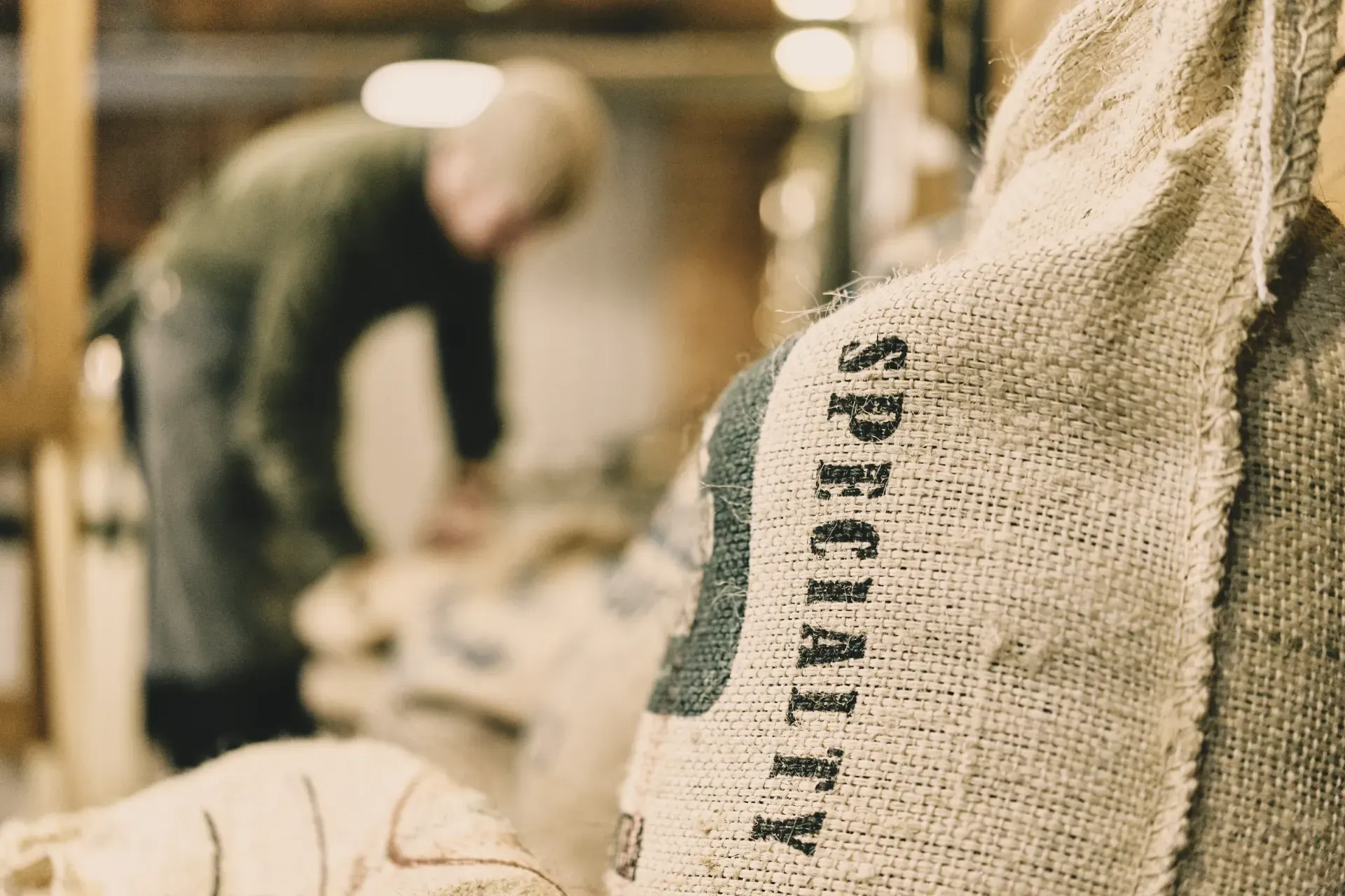 A burlap sack stamped with the word SPECIALTY fills the foreground in sharp focus. In the background, a blurred figure of a worker bends over their work, unnamed and secondary to the label.
