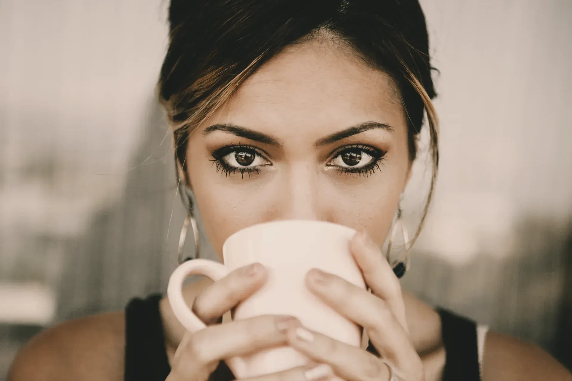 A close-up portrait of a woman holding a coffee cup over her mouth and lower face. Her eyes look directly into the camera — present, aware, unheard.