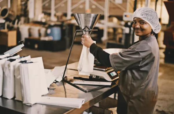 A woman in a hairnet and work apron fills coffee bags at a packaging station in a roastery. She is smiling and looking toward the camera 