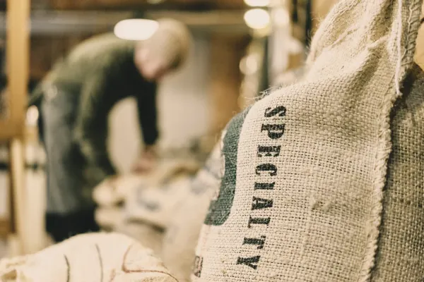 A burlap sack stamped with the word SPECIALTY fills the foreground in sharp focus. In the background, a blurred figure of a worker bends over their work, unnamed and secondary to the label.