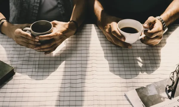 Two people sitting across from each other at a checkered table, each holding a cup of coffee, seen from above.