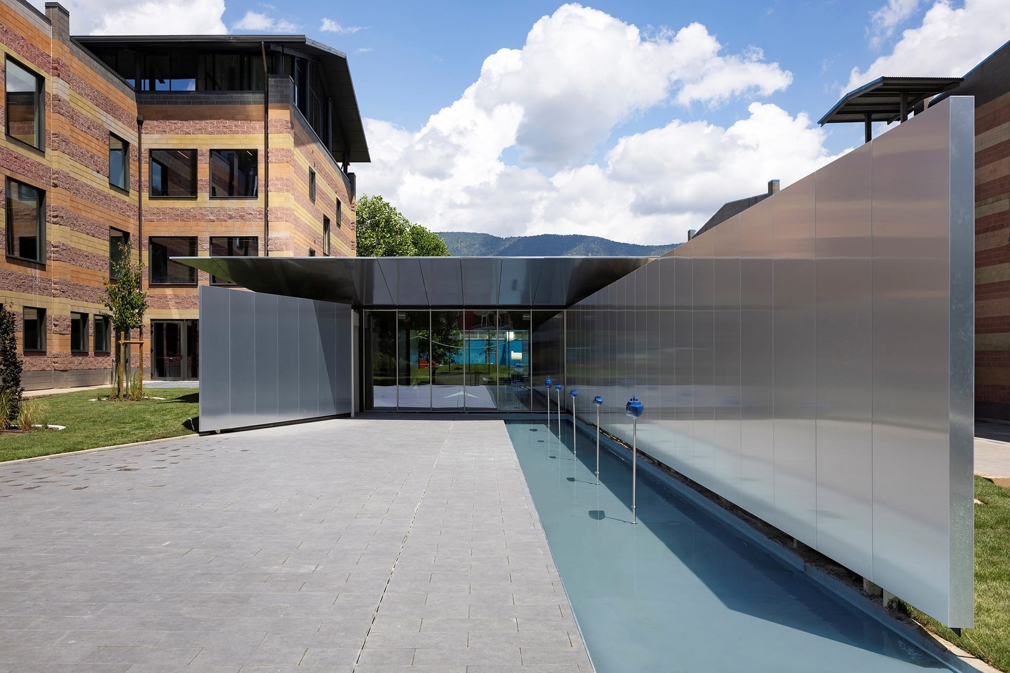 Eingangsbereich des Azimut-Hauptsitzes in Avigliana bei Turin mit einem Klinkerbau, einer Wand aus gebürstetem Metall, einem überdachten Glaseingang und einem langen Wasserbecken mit kleinen blauen Bootsmodellen.