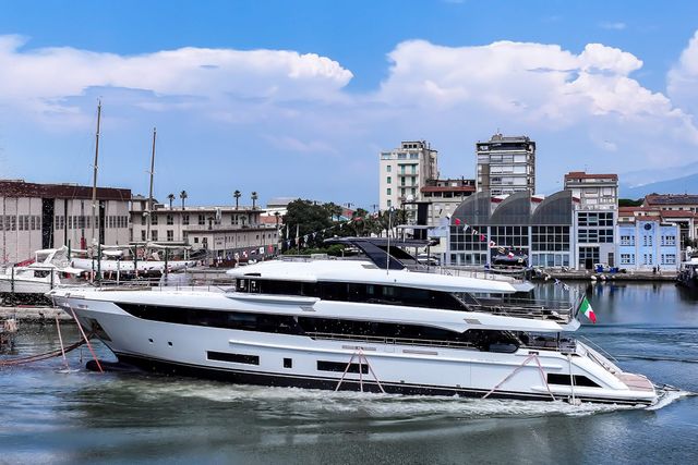 Die weiße Superyacht Benetti Class 44M „Lumina“ gleitet beim Stapellauf von der Slipanlage ins Hafenbecken von Viareggio, über ihr gespannte Signalflaggen, im Hintergrund Werfthallen und Wohn