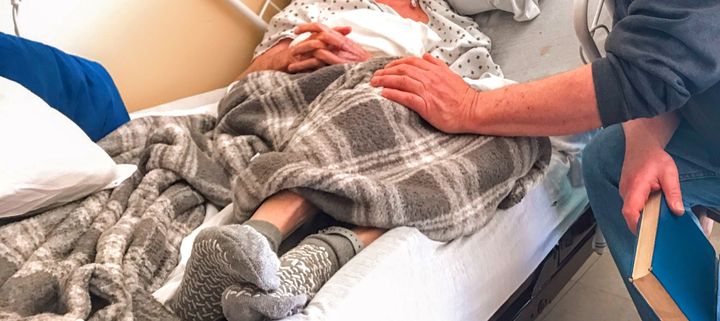 One man visiting another in a hospital bed. He holds a book and touches the other gently on the knee.