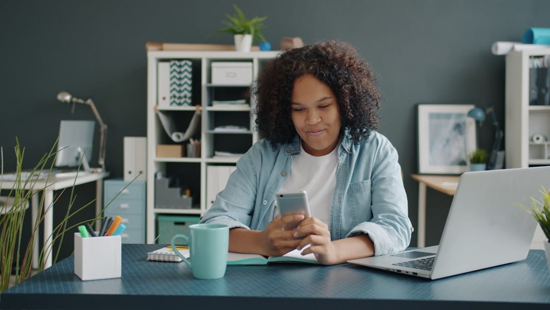 Young woman using smartphone at desk with laptop.