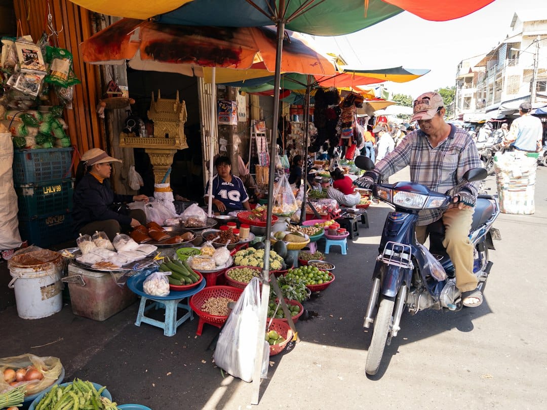 A person on a motorbike passing a street market with colorful umbrellas and produce