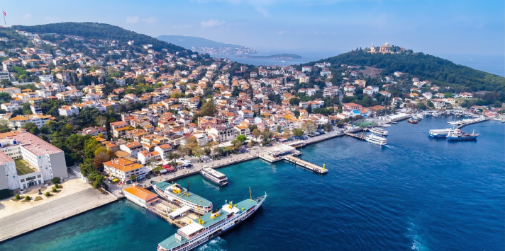 İstanbul Prens Adaları'ndan Heybeliada'nın havadan kuş bakışı görünümü; kıyıdaki vapur iskelesi, yanaşmış Şehir Hatları vapuru, turkuaz deniz ve tepedeki tarihi konaklar.