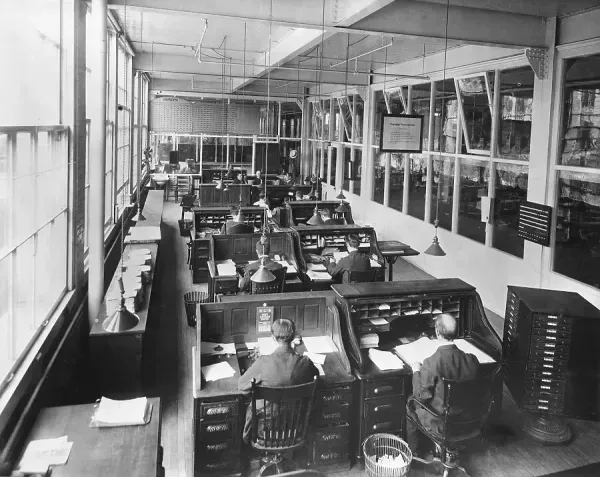Early twentieth-century open office with clerks and supervisors, illustrating apprenticeship through proximity rather than formal credentials.