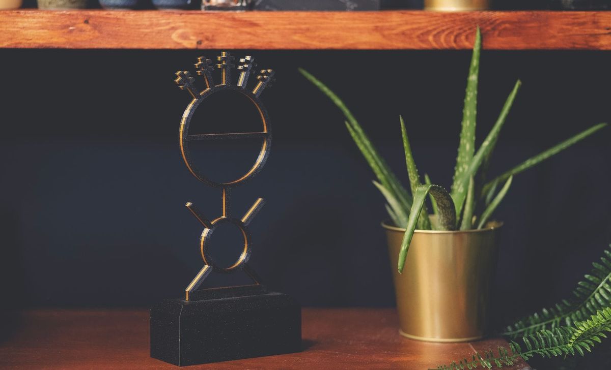 Bob Proctor Stick Man metal sculpture on a wooden shelf beside a potted aloe plant.