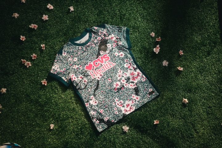 A photo of the new Washington Spirit jersey. It is mostly dark green, speckled with white, with white cherry blossoms scattered throughout the print. The collar, cuffs, and ribs are solid da