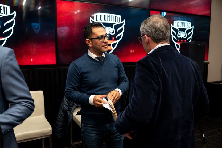 A photo of D.C. United managing director of soccer operations Dr. Erkut Sogut speaking with a man in front of screens featuring the D.C. United logo.