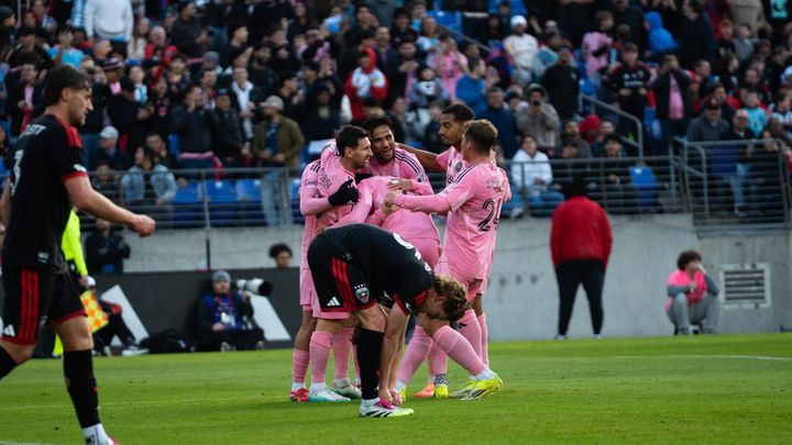A photo of Inter Miami players celebrating a goal while D.C. United's Kye Rowles leans down to re-tie his shoes.