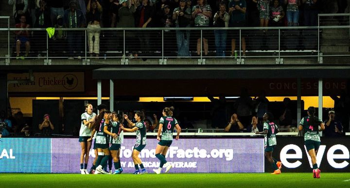 Several Washington Spirit players celebrate a goal at Audi Field, with fans in the background also celebrating.