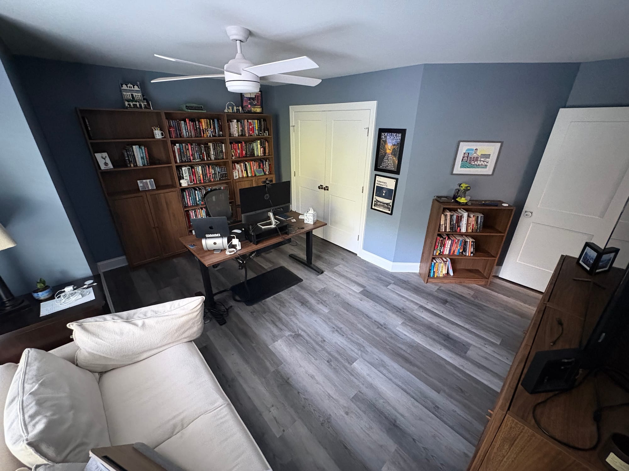 An office picture taken from the corner of the room. In the middle of the room, an espresso standing desk. Behind it, three tall Ikea Billy Bookshelves lined with books, kids art work, and lego sets. On the wall, a poster with Philip's name on it from his Noir at the Bar performance in 2019 and a Tottenham Hotspur poster.