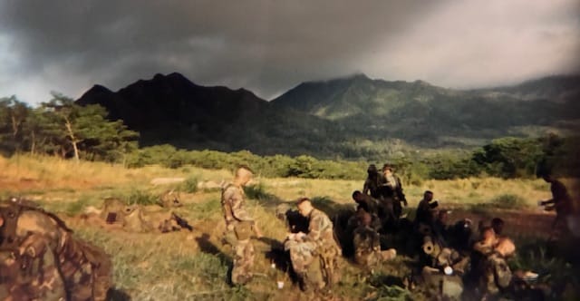 An Infantry squad sits in a green field, dark green, volcanic mountains are shrouded in dark clouds in the background