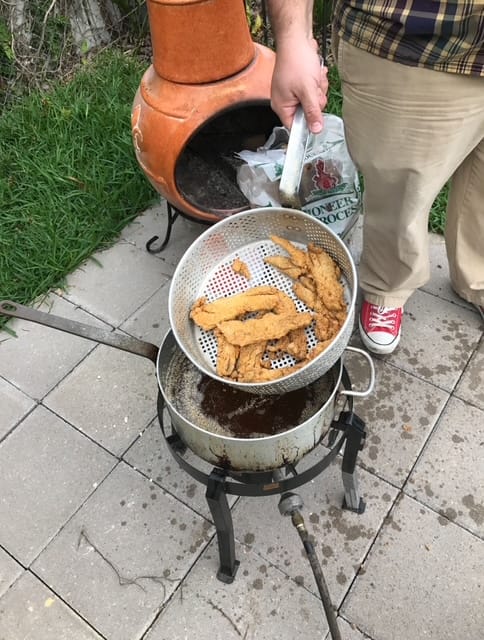 A fish cooker and pot of oil on a propane burner.  An unknown male holds up a basket of perfectly fried fish fillets.