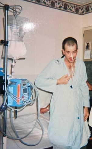 Patient in blue hospital pajamas standing next to a bed with an IV pole and a feeding tube.
