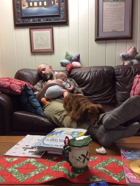 A white male sleeps on a couch with a dog and an Infant asleep on his legs and chest.