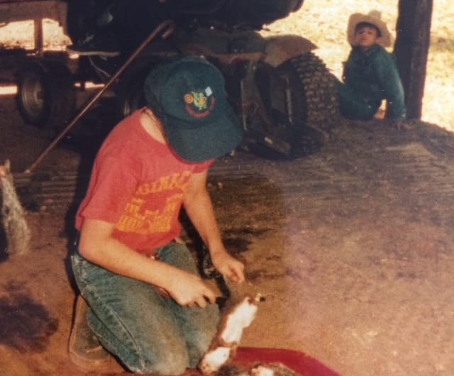 Grainy photo.  A young boy in a blue shirt and a cowboy hat watches another in a red shirt clean a Squirrel.