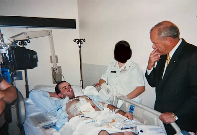 A patient in his mid 20s lays in a hospital bed covered in bandages and apparatus while an anonymous nurse and a white man in a suit look on.