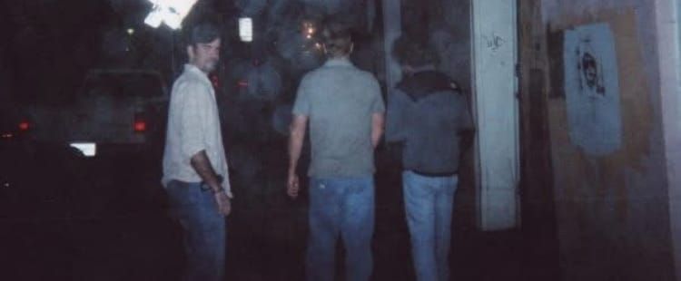 Three young men walk down a fog shrouded night street.  The one on the far left is looking back at the camera.