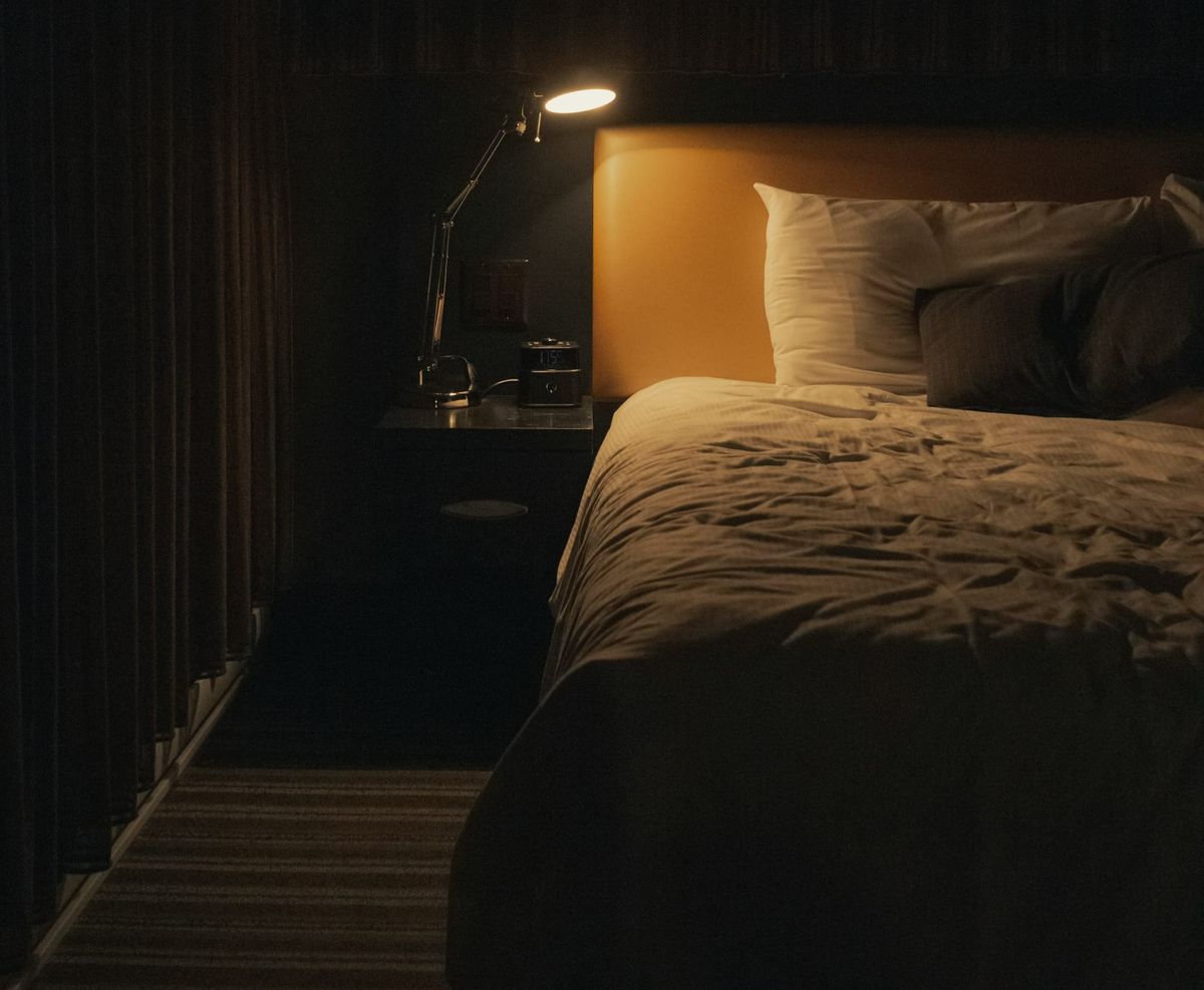 A bedside lamp shins dim light on an empty bed in an otherwise dark bedroom