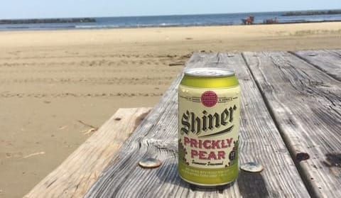 A can of Shiner Prickly Pear beer sitting on a battered picnic table on a beach overlooking the gulf of Mexico