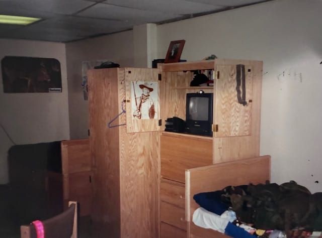 A dingy barracks room with beige walls, tan furniture and broken, stained, white ceiling tiles.