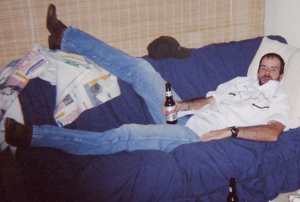 A bearded white man in white shirt and jeans laying on a couch covered with a blue blanket drinking a Coors Light long neck.