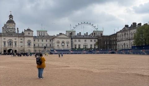 A well manicured brown dirt drill field is in the foreground.  A row of old English buildings in the background.  The London Eye ferris wheel is in the far distance against.