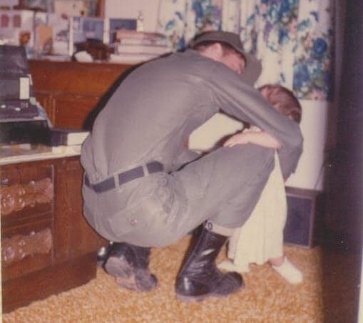 A soldier in green uniform kneels to hug a small child.