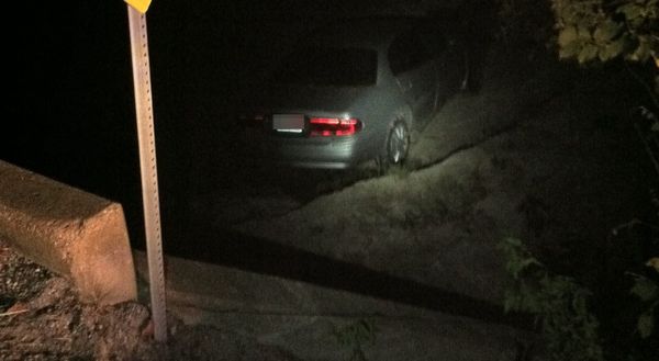 A night time photo of a beige car stuck deep in a roadside ditch.