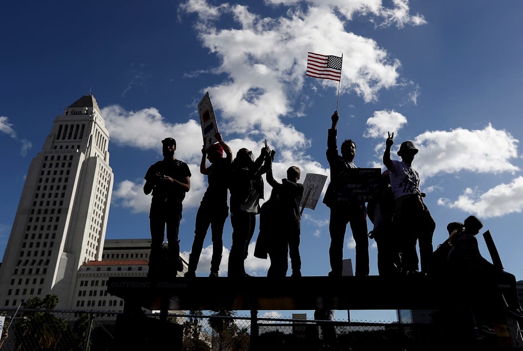 在美國加洲洛杉磯，有抗議者爬上巴士站上高舉標語牌。