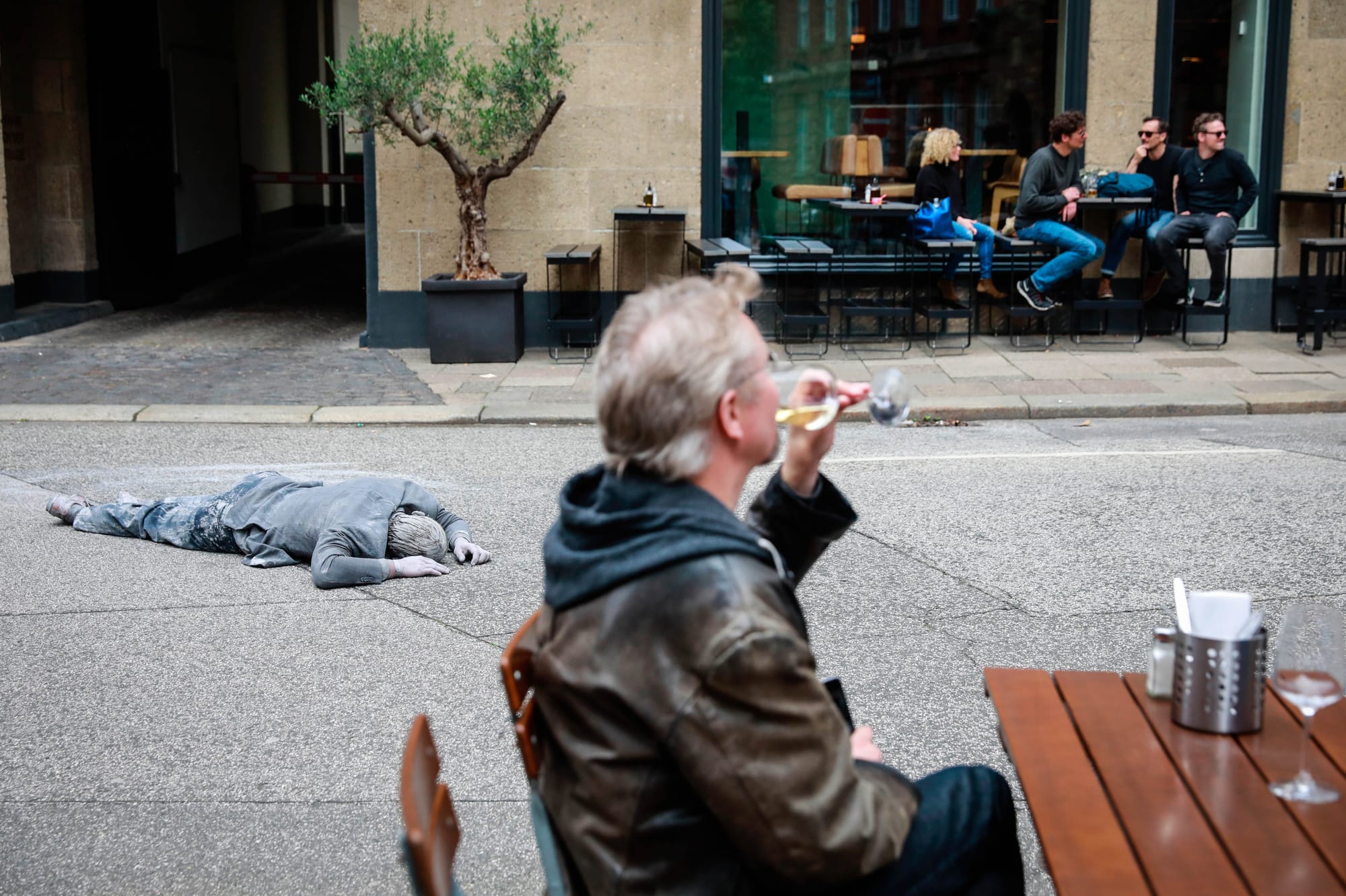 2017年7月5日，一名「1000 Figures」的參與者，在漢堡街頭的戶外酒吧前，躺在街道上，彷如死人。旁邊的酒客仍品嘗著杯中美酒。