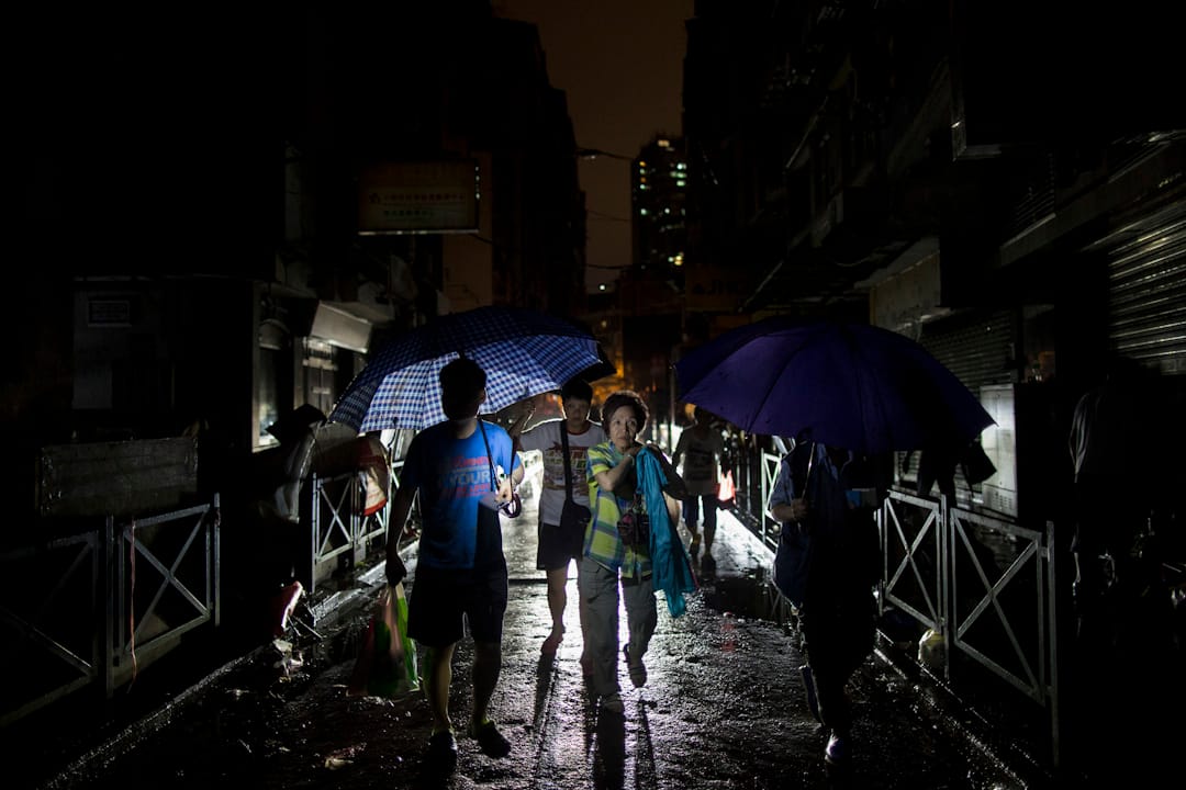 入夜后，澳门的街道仍然漆黑，还下起雨来，市民撑著伞，靠走过的车辆的车头灯照明，横越街道。