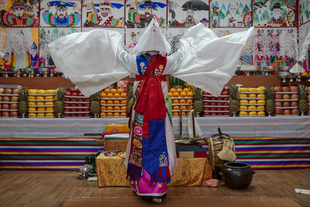 韩国始兴市，萨满巫师Nunkotsunnyu正在进行“跳神”仪式，教徒相信仪式能够带来运气，荣誉，健康及避免邪灵带来灾害。墙壁上贴满神灵的画作及护符避免邪灵干扰仪式。