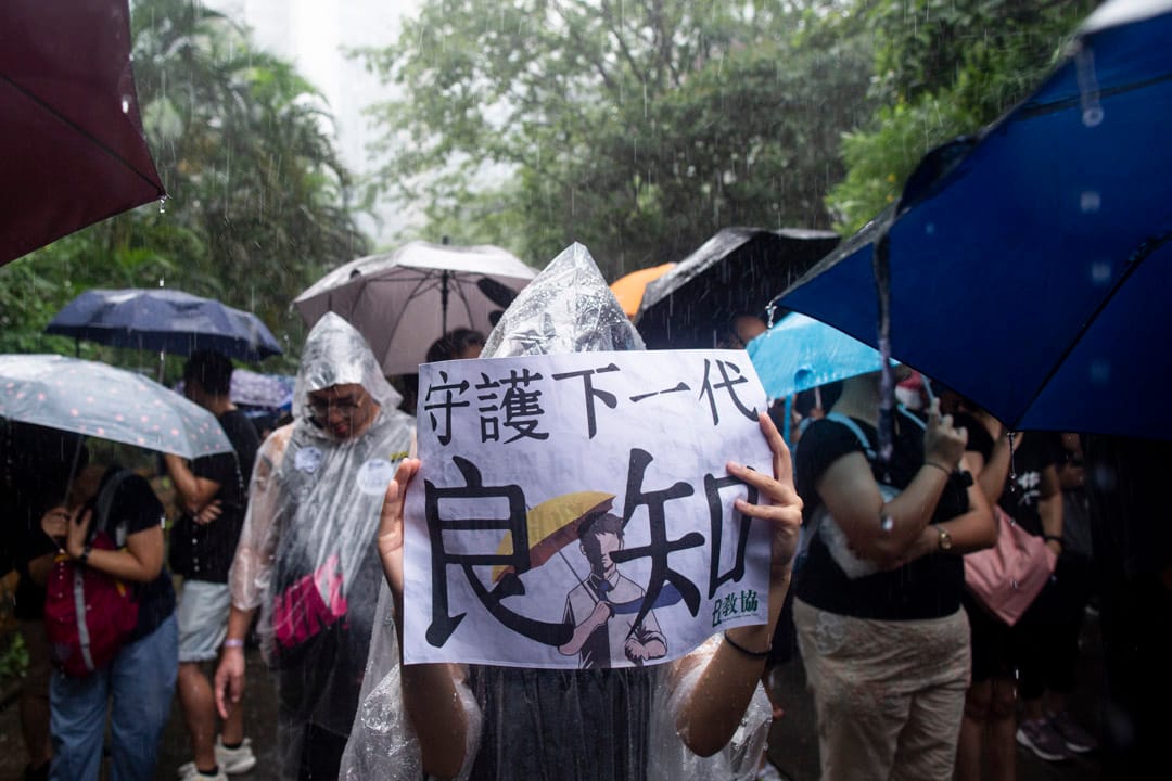 2019年8月17日，有教师发起“守护下一代，为良知发声”的游行，期间大雨滂沱，参加者身上贴上“我是香港教师”的贴纸，又手持“守护下一代良知”、“凭老师守护”的标语。 摄：林振东/端传媒 