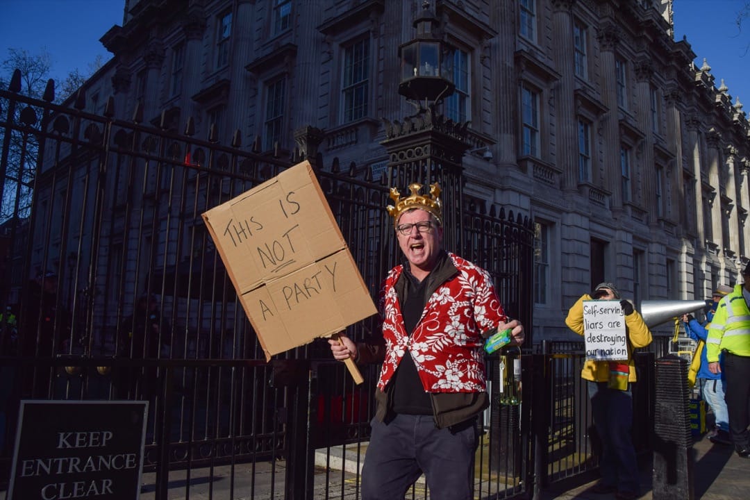 2022年1月12日，英國首都倫敦，一名示威者手持標語、芝士和白酒，到首相府門外示威，要求首相約翰遜就「派對門」事件問責。