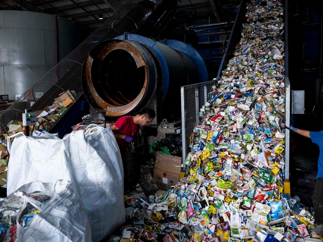  紙漿機器入口處有兩條運輸帶，一條佈滿紙包飲品盒，一條堆積雜廢紙，