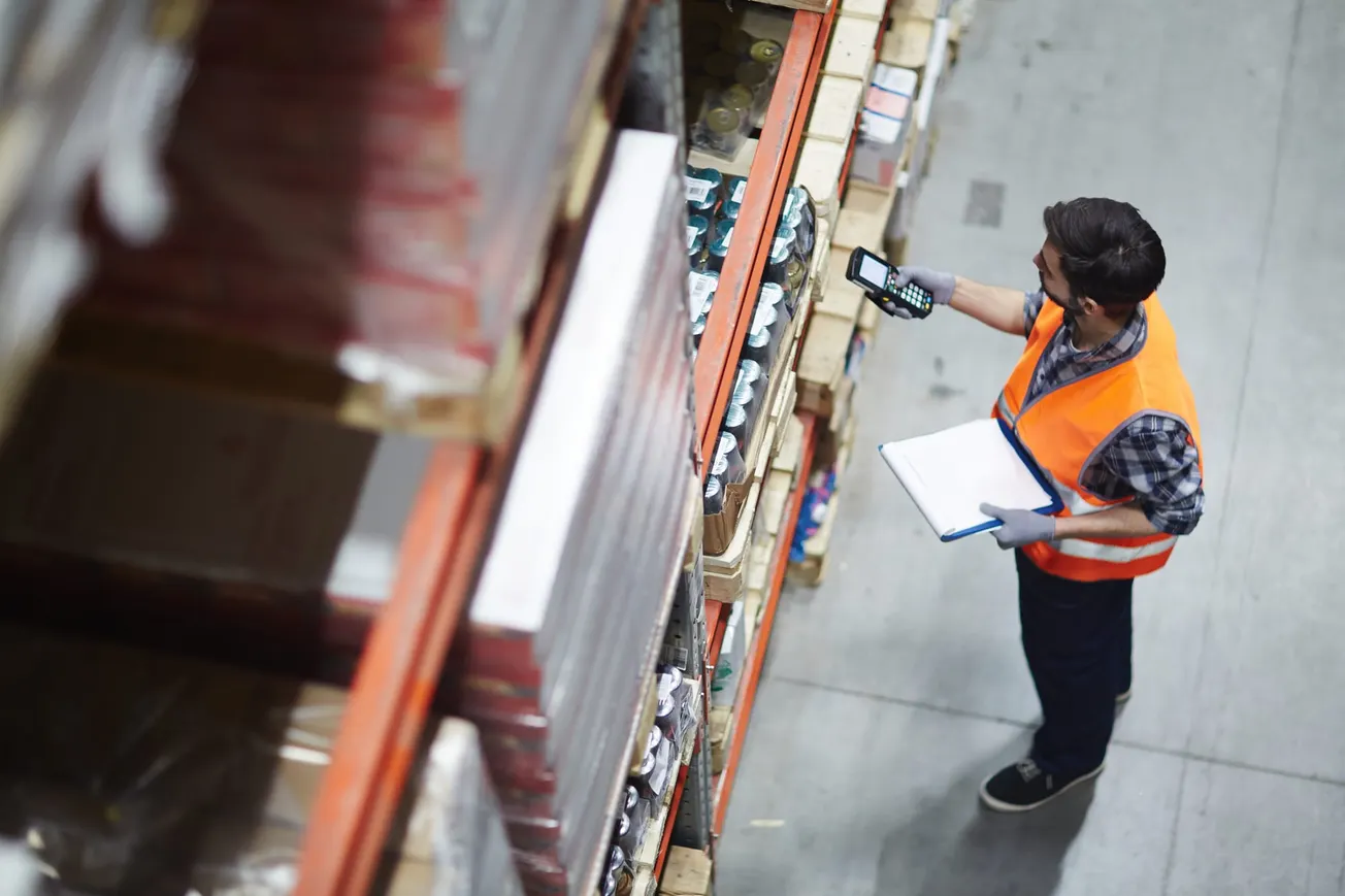 Warehouse worker using a handheld scanner and clipboard to manage inventory on shelves, representing supply chain operations and logistics management.