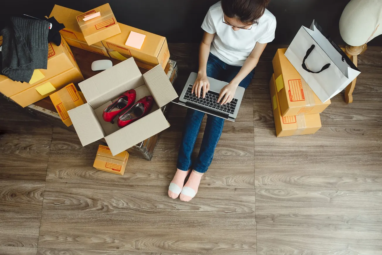 Woman managing online retail business from home, surrounded by shipping boxes and using a laptop to fulfill e-commerce orders.