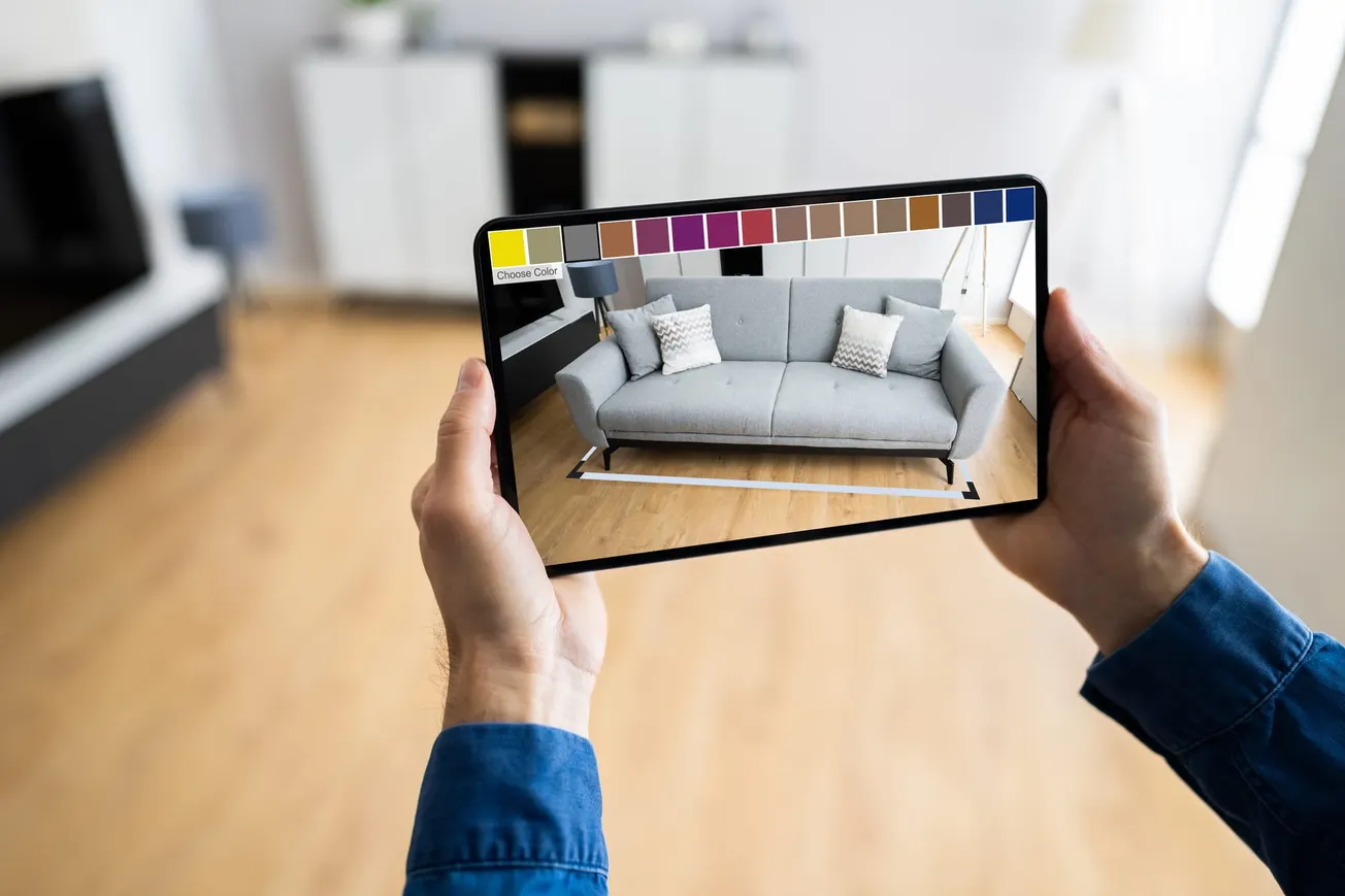 Person using a tablet to visualize a gray sofa with augmented reality (AR) furniture app in a modern living room.