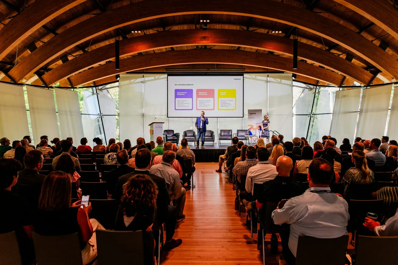 Spacious conference hall with a curved wooden ceiling, a speaker on stage, and an audience seated attentively. Presentation slide brightly lit.