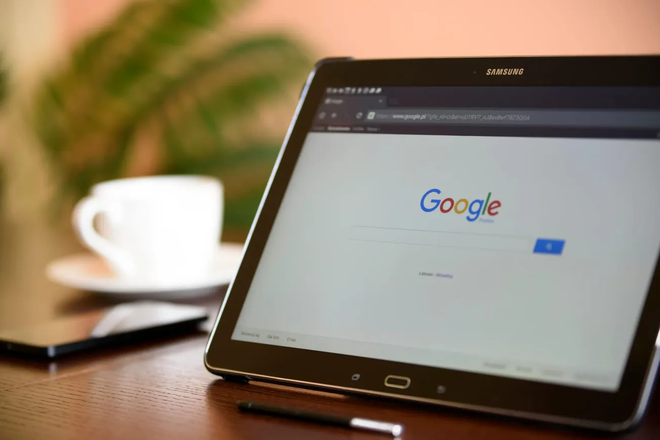 Tablet with Google Search open on a browser, placed on a wooden table near a phone and a white cup.