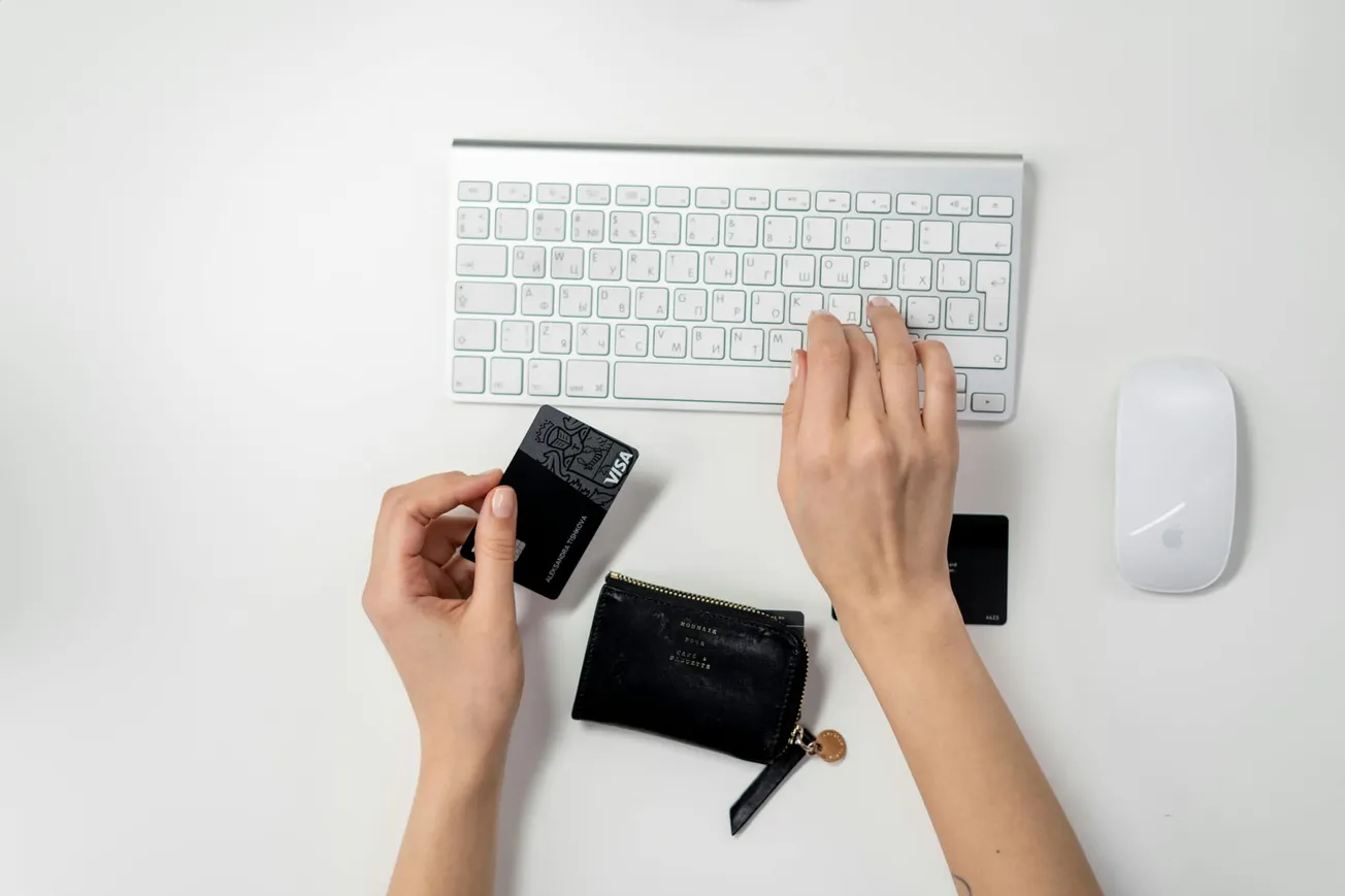Hands using a computer keyboard and mouse, with a black credit card in one hand and a small black wallet nearby, indicating online shopping.