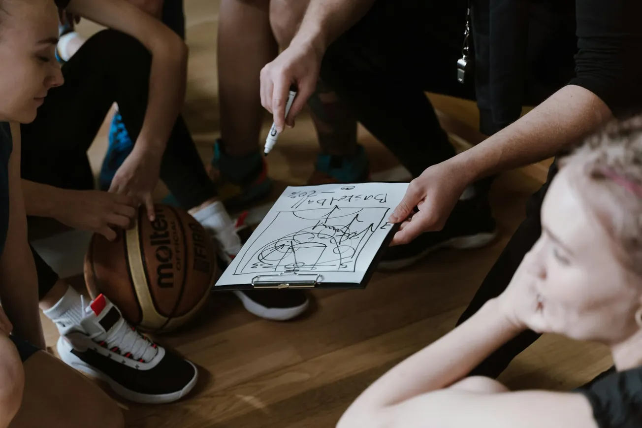 Coach holds clipboard with basketball play as team huddles on court. Players listen attentively, one holding a ball.